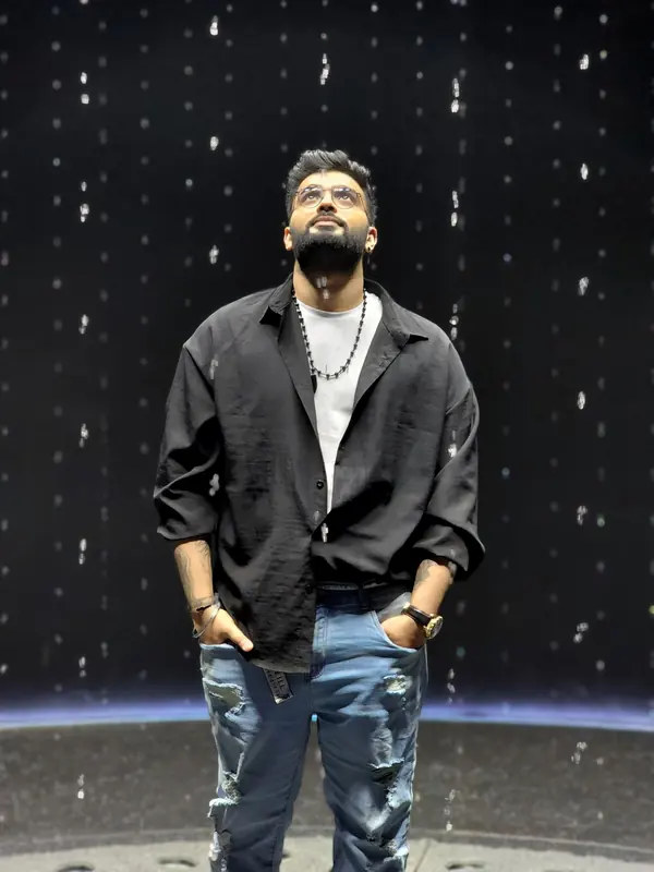 Portrait of Just AMN, open-format DJ, standing in a black shirt and ripped jeans against a dark backdrop